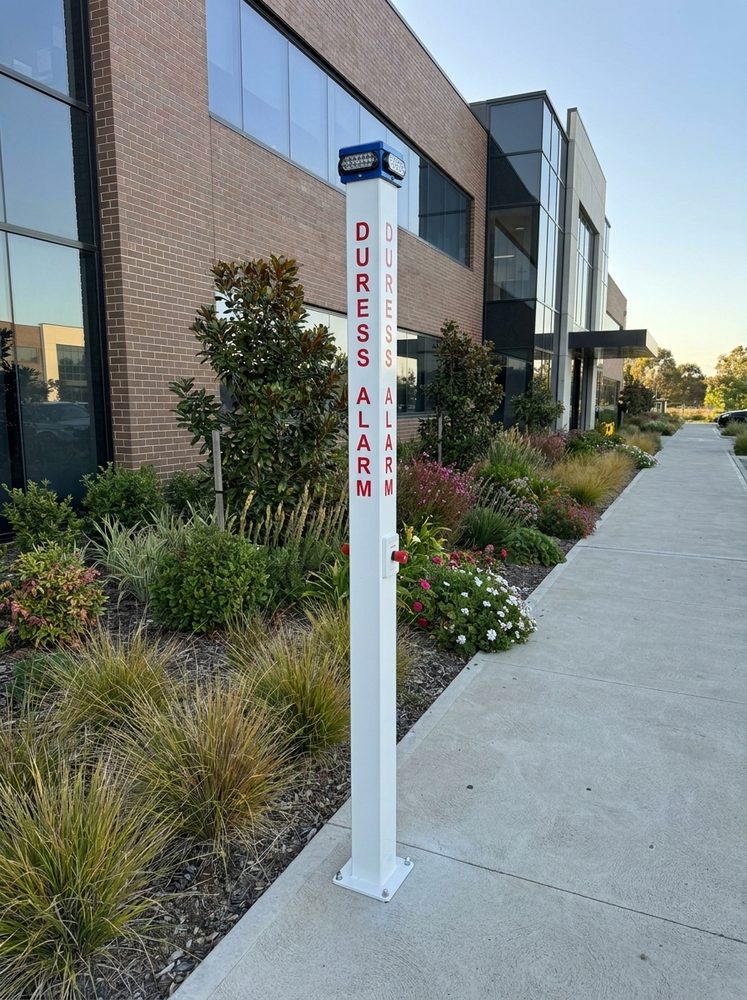 A white duress alarm pole with a blue LED beacon head, installed on a footpath beside a commercial building.